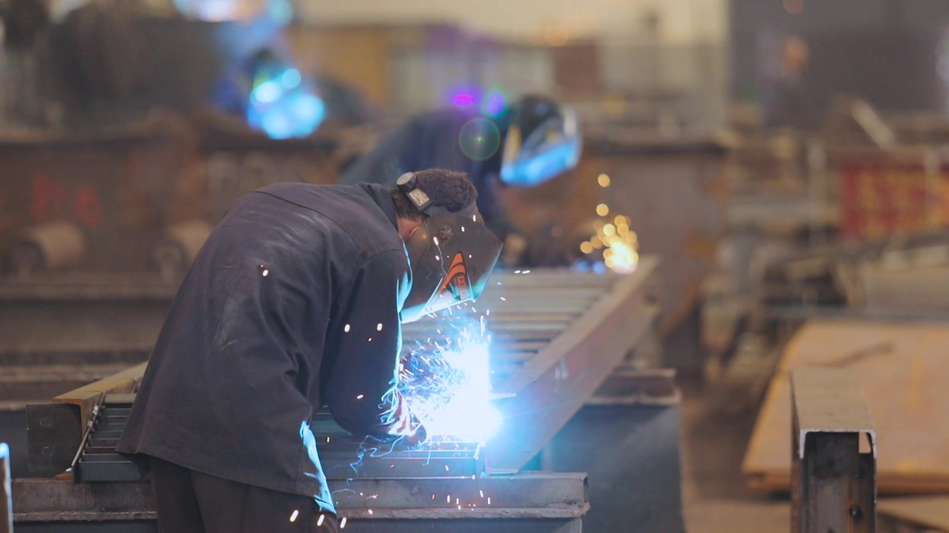 a welder wearing a welding mask in a fabrication facility working on a steel beam. bright blue sparks are flying